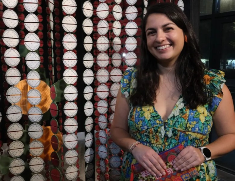 A smiling woman with long dark hair stands next to a hanging art installation composed of vertical strands of white and red circular discs. She is wearing a sleeveless, floor-length dress with a blue, green, and yellow citrus fruit print and holding a colorful embroidered clutch.