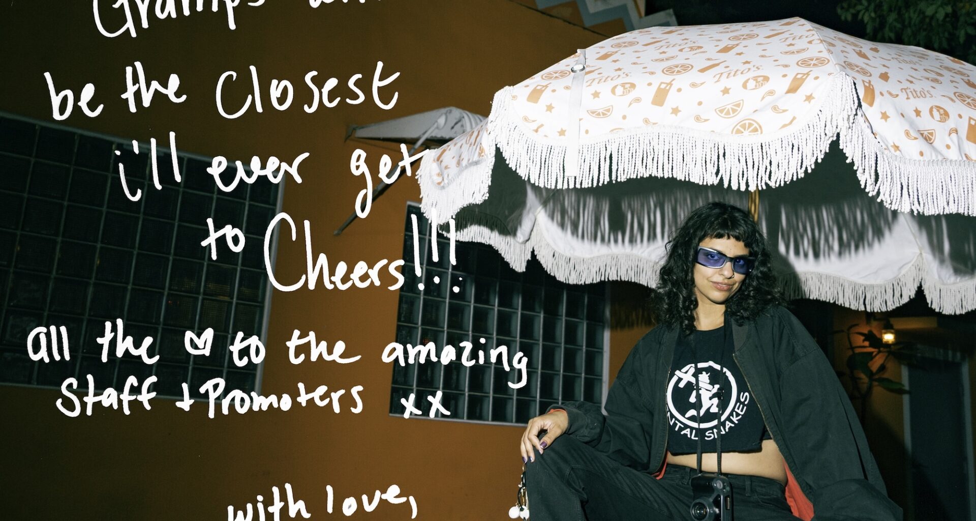 Photo of a woman sitting under an outdoor restaurant umbrella in front of an orange building. Text written on the photo reads, "Gramps will be the closest I'll ever get to Cheers! All the [heart symbol] to the amazing staff + promoters xx. With Love, Amanda"