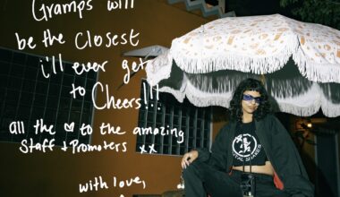 Photo of a woman sitting under an outdoor restaurant umbrella in front of an orange building. Text written on the photo reads, "Gramps will be the closest I'll ever get to Cheers! All the [heart symbol] to the amazing staff + promoters xx. With Love, Amanda"