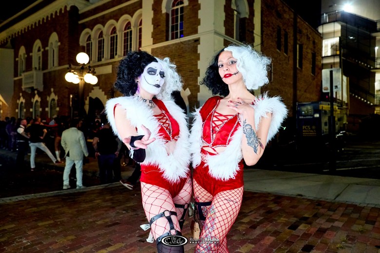 A promotional photo of two performers outdoors at night, standing in front of an historic red brick building. The subjects wear high-contrast outfits with black and white hair, red lips, and dramatic makeup.