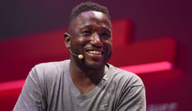 A photo of a Black man in a gray t-shirt smiling while wearing a headset microphone.