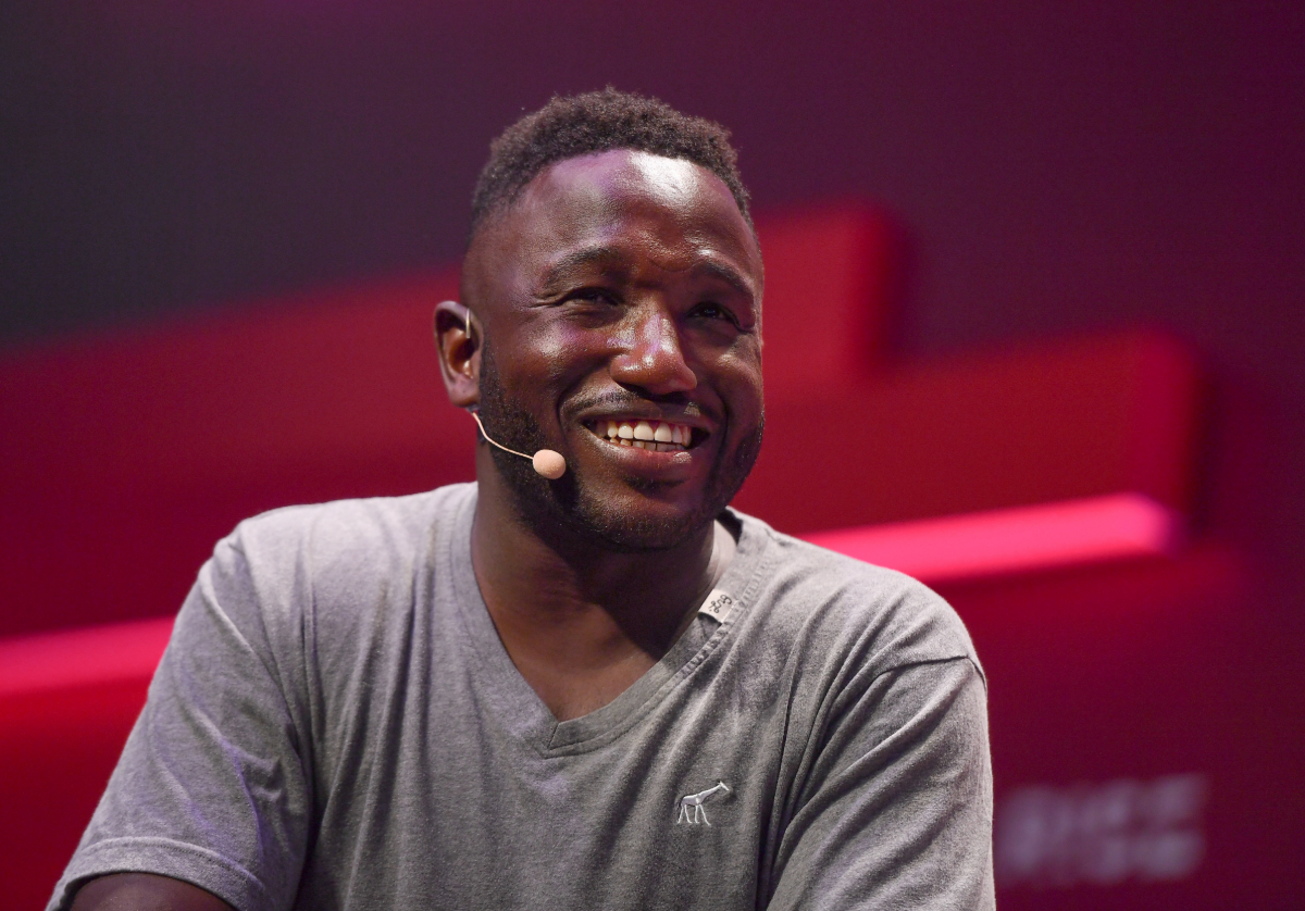 A photo of a Black man in a gray t-shirt smiling while wearing a headset microphone.