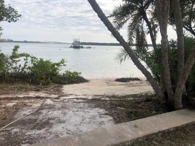 view from behind a low concrete seawall looking out over a small sandy beach toward a calm bay. A large boat is anchored in the distance under a cloudy sky.
