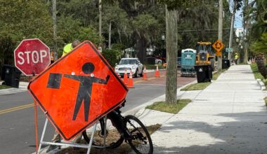 an orange construction sign cautions there is a flag man ahead, as he holds a red stop sign with a construction project ongoing on a side street.