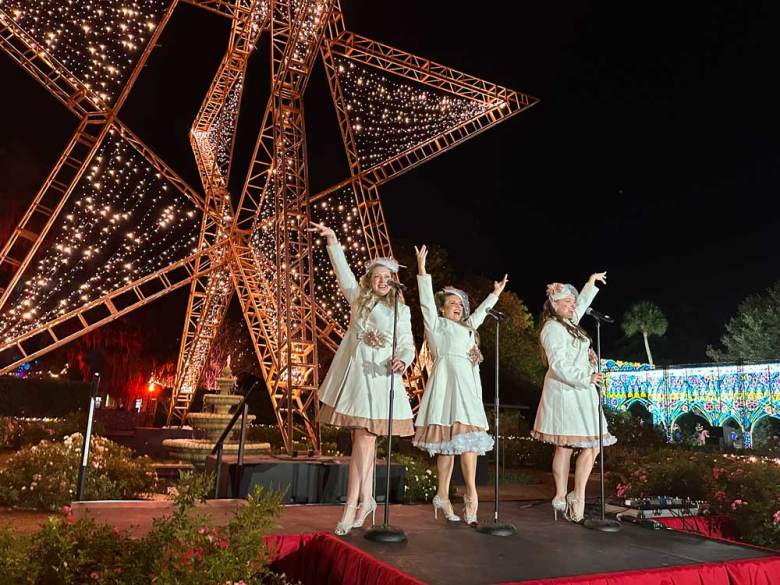 a trio of women dressed in white singing under a large star made of twinkle lights