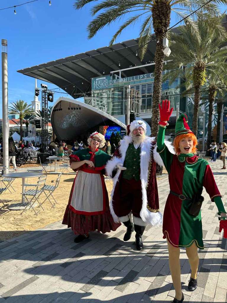 Mrs. Claus, Santa Claus, and a redheaded elf in front of the Dr. Phillips Center for the Arts
