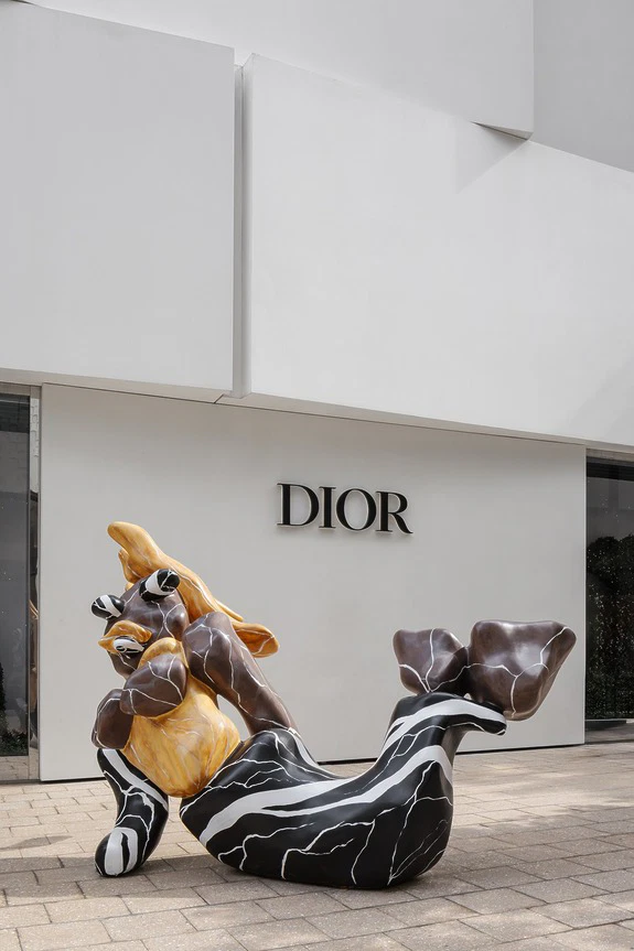 Abstract sculpture resembling a reclining figure in front of Dior store with a modern white facade.