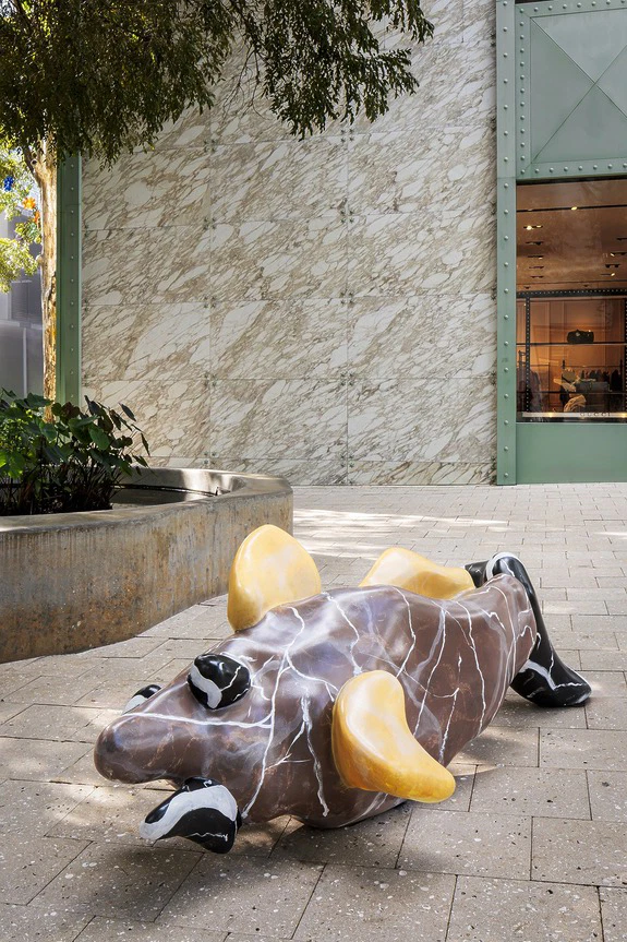 Sculpture of a playful, abstract animal with yellow fins and marble texture lying upside down outside a modern building.