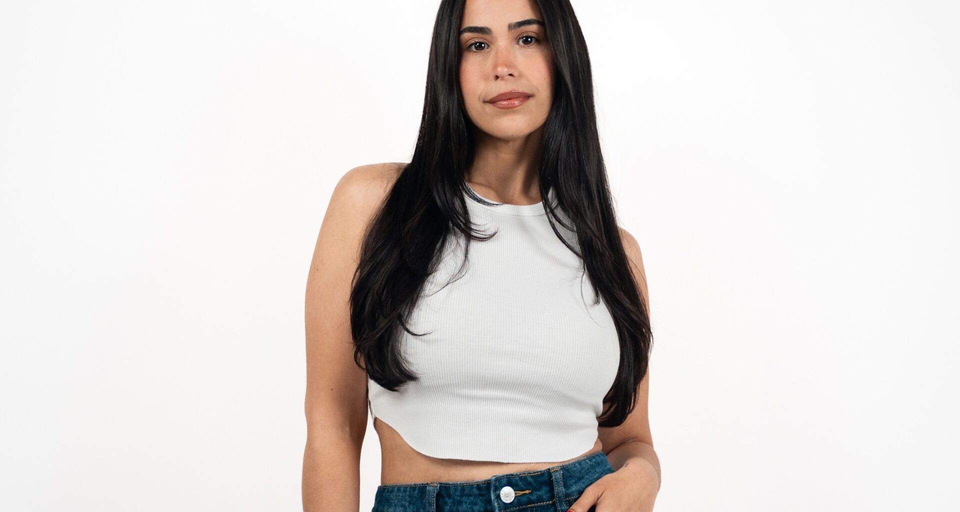 photo of a woman with long black hair in a white crop top and jeans posing in front of a white backdrop