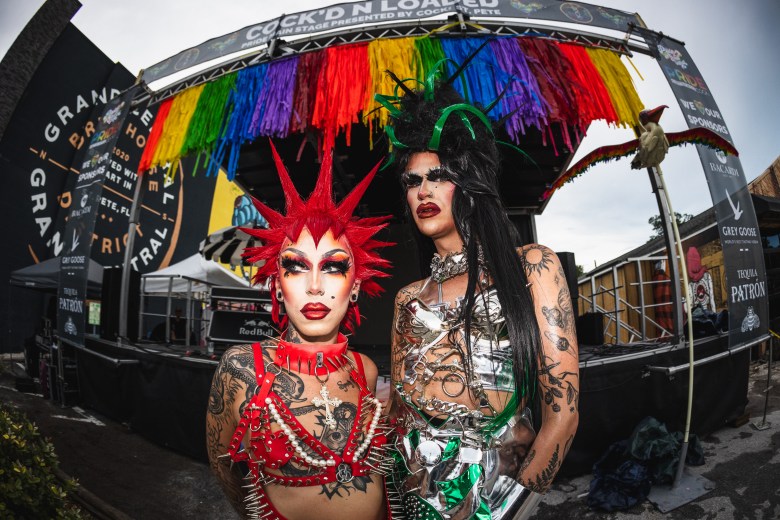 Two drag performers pose in front of an outdoor stage decorated with rainbow fringe. The performer on the left features a bright red spiked wig and a red harness adorned with pearls and spikes, while the performer on the right wears a metallic silver outfit and a tall black wig with green accents. Banners for "Grand Central" and "Cock'd N Loaded" are visible on the stage structure behind them.