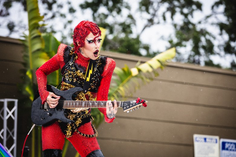A performer with elaborate red hair and dramatic makeup plays a black electric guitar on an outdoor stage in the rain. They are wearing a red, crystal-encrusted bodysuit with gold detailing and a black harness featuring a lightning bolt strap. Raindrops are visible against the background of trees and a building, where a "POOL RULES" sign can be seen in the lower right corner.