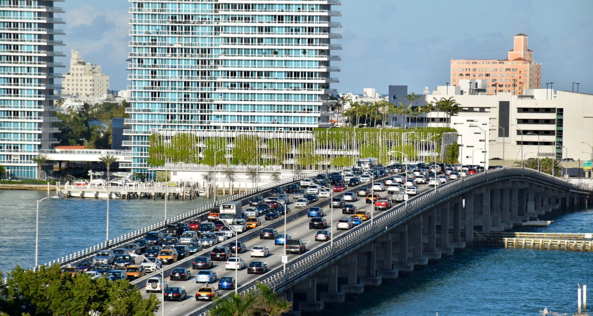 three lanes of cars sit in standstill traffic on a bridge over blue water, with high-rise towers in the background