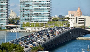 three lanes of cars sit in standstill traffic on a bridge over blue water, with high-rise towers in the background