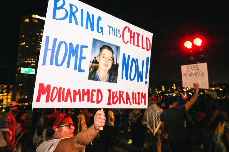 Nighttime protest scene where a woman holds a large sign that reads "BRING THIS CHILD HOME NOW! MOHAMMED IBRAHIM" with a photo of the boy in the center. Other protesters and city lights are visible in the background.