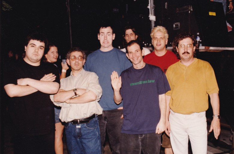 A candid-style portrait against a mottled brown background. An individual in a black t-shirt embraces a dog, who is looking away from the camera. The lighting is soft and golden, creating a gentle and affectionate atmosphere.
