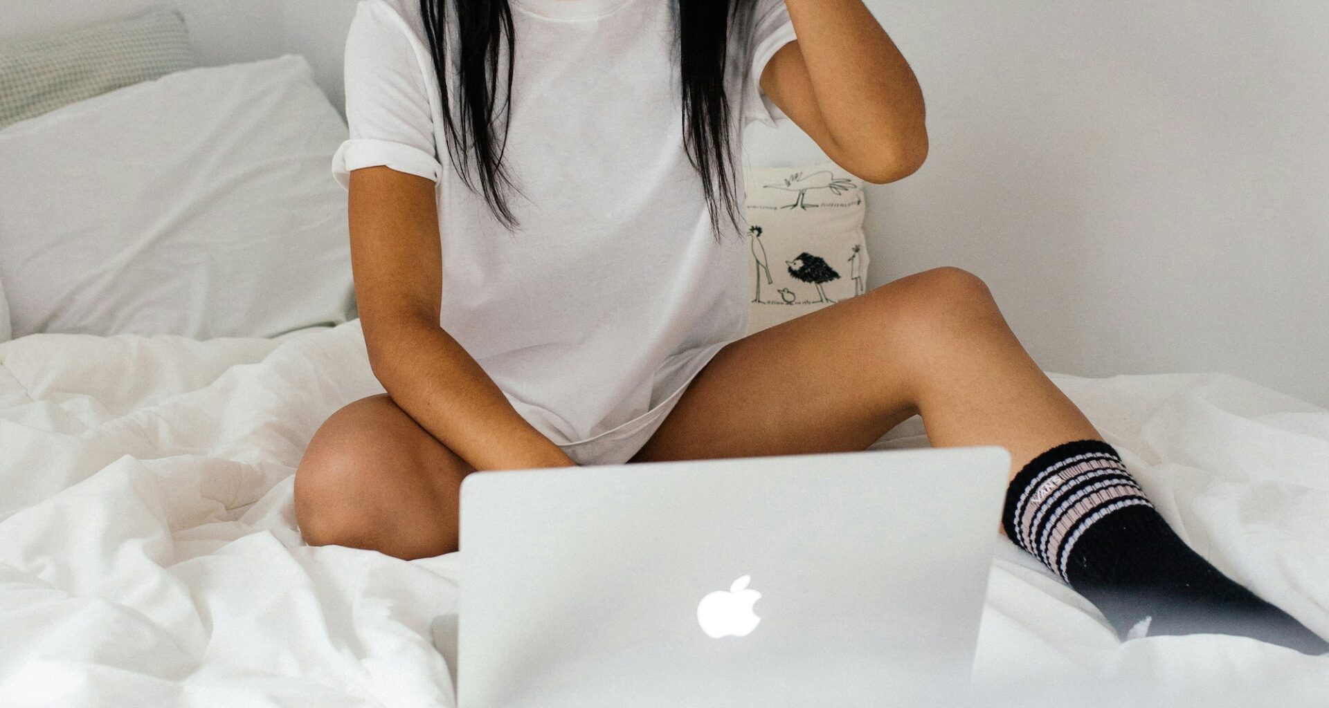 photo of a woman in a t-shirt and socks in front of a laptop on a bed