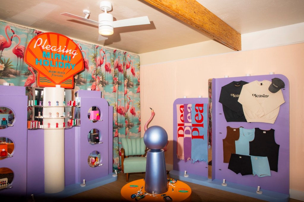 Photo of a room with a sign shaped like a seashell reading, "Pleasing Miami Holiday" with purple and beige shelves holding nail polish, scarves, and T-shirts. A curtain with flamingos hangs on the back wall.