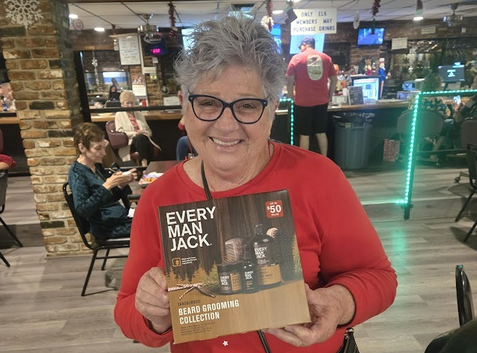 A smiling older woman with short grey hair and black-rimmed glasses stands indoors, holding up a boxed "Every Man Jack" Beard Grooming Collection. She is wearing a red long-sleeved shirt and a black crossbody bag. The background shows a casual social hall with brick columns, holiday decorations, seated guests, and a sign above the bar that reads "Only Elk Members May Purchase Drinks."
