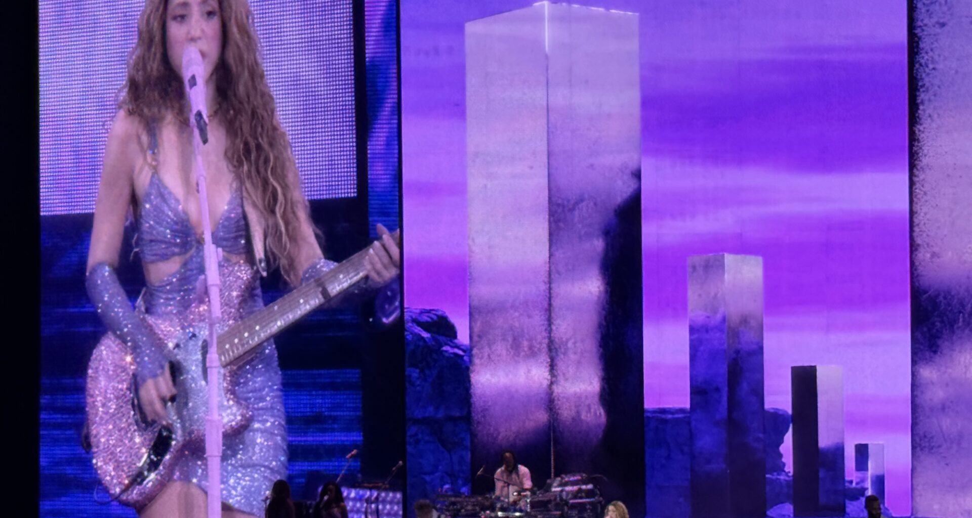 photo of Shakira singing and playing guitar onstage with a Jumbotron projecting her image behind her