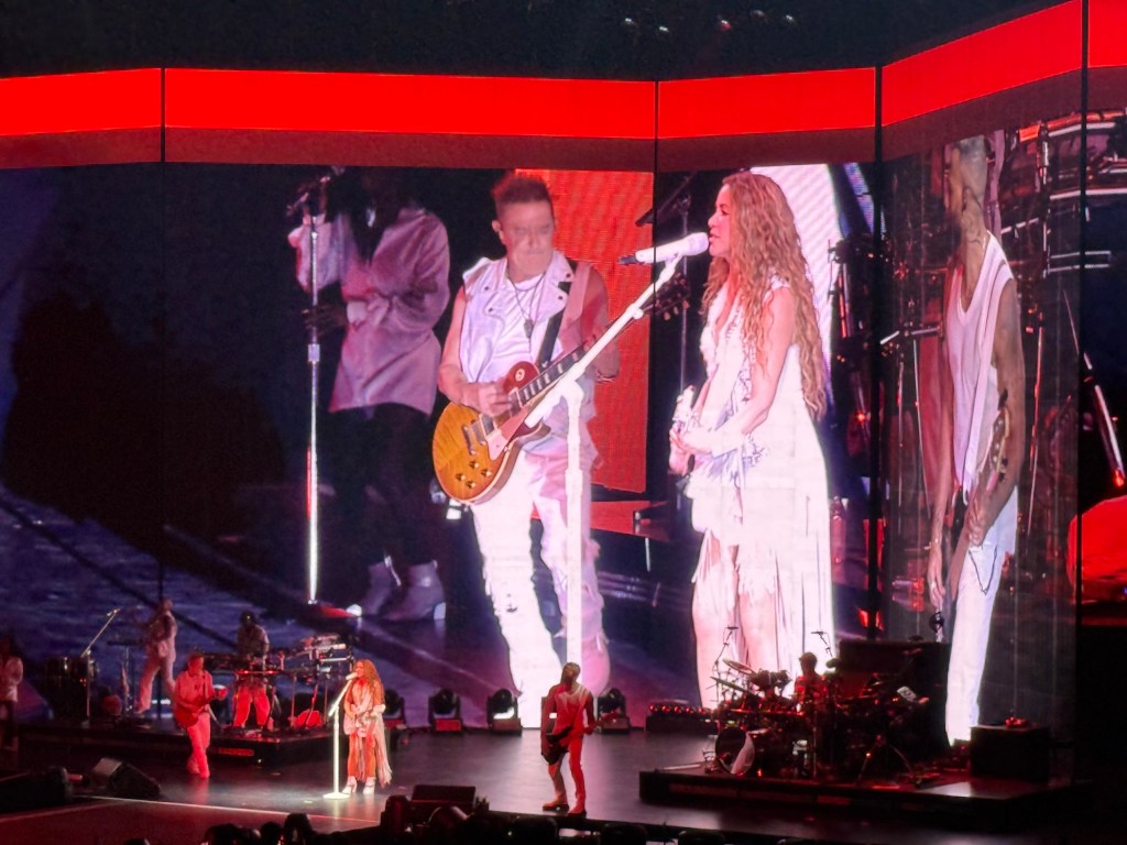 photo of Shakira singing and playing guitar onstage with a Jumbotron projecting her image behind her