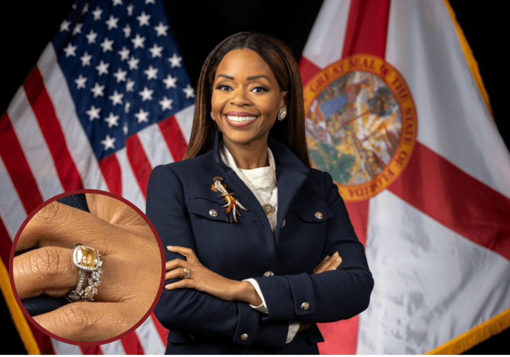 A Black woman stands with her arms folded in front of flags for the State of Florida and United States of America.