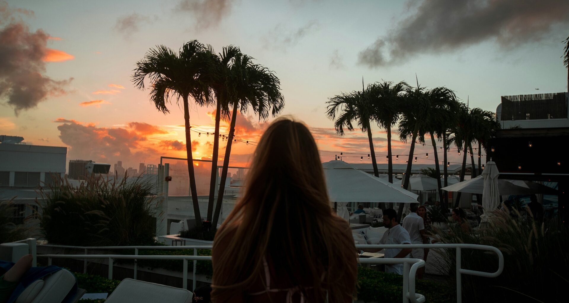 photo of the silhouette of a women watching the sun set behind palm trees on a hotel rooftop