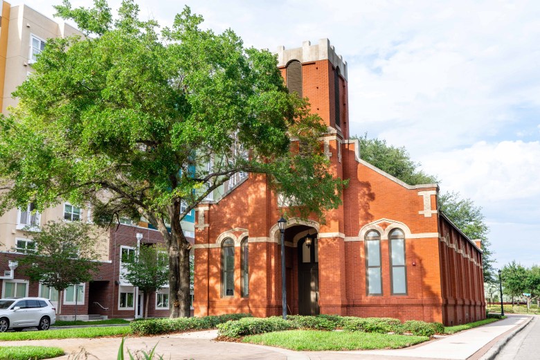 The historic St. James Church, a red brick structure, is seen on a sunny day in downtown Tampa, situated on a grassy corner lot adjacent to a new apartment complex and a paved sidewalk.