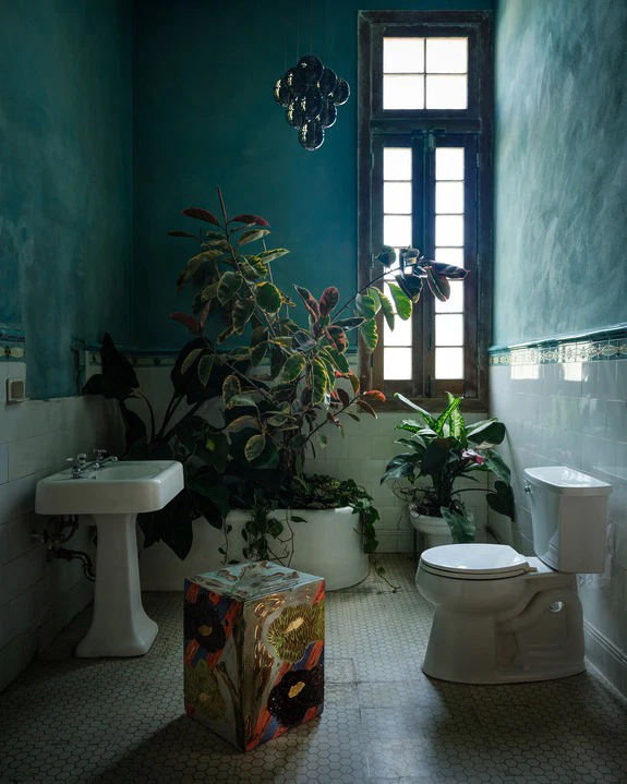 Bathroom with teal walls, white fixtures, large window, and lush green plants arranged around the room for a natural touch.