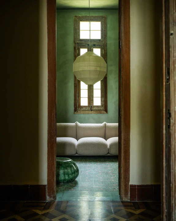 Cozy living room with a plush beige sofa, green walls, large paper lantern, and vintage tiled floor viewed through a doorway.