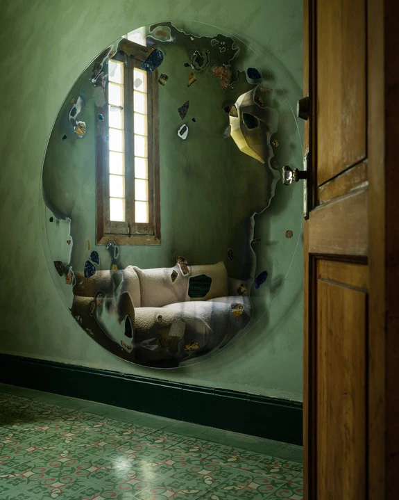 Large circular decorative mirror on a green wall reflecting an open wooden door and a patterned floor in a bright room