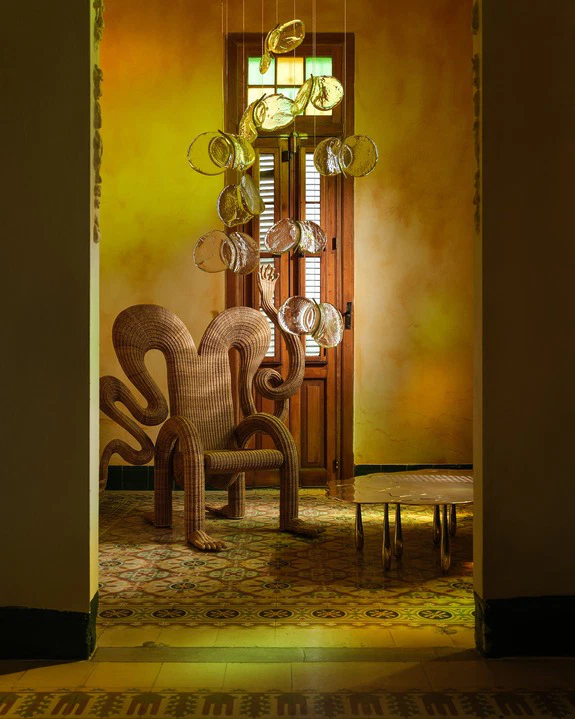 Wicker chair with unique design and artistic lamp in sunlit room with patterned tiles and wooden door background.