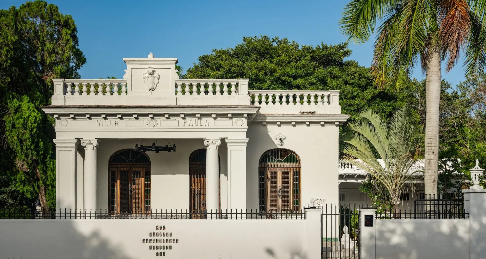 Historic white villa with arched windows and ornate detailing, surrounded by lush greenery and a palm tree.