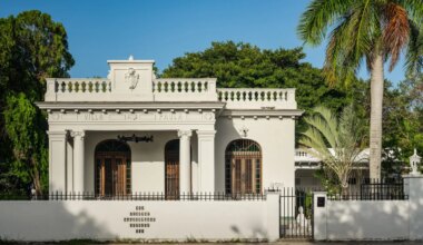 Historic white villa with arched windows and ornate detailing, surrounded by lush greenery and a palm tree.