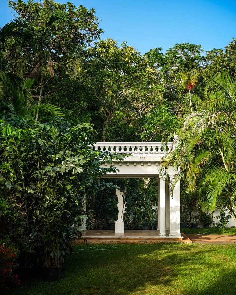 White garden gazebo surrounded by lush green trees and plants under a clear blue sky.