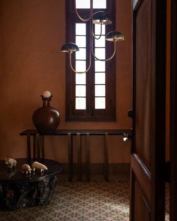 Dimly lit room with a modern chandelier, wooden door, decorative table with sculptures, and a window with natural light.
