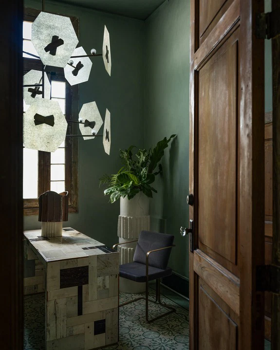 Room with wooden desk, modern hanging light fixture, green walls, potted plant, and a single chair near an open door.