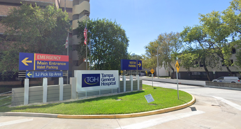 A clear, sunny photo of the Tampa General Hospital entry drive, showing multiple directional signs and the TGH logo sign set in a grassy median in front of the multi-story hospital building.