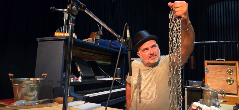 An individual, identified as Juan Rodriguez, performing as a Foley artist on stage. The person, wearing a fedora and a graphic t-shirt, holds up a heavy metal chain in the air while looking towards it. A piano, a microphone, and various sound props are set up around them.