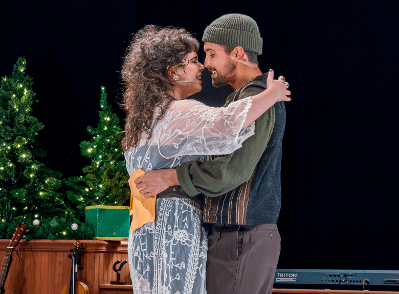 Two performers embracing on a dark stage lit by warm spotlights, likely from a holiday production. The person on the left has long, curly hair and wears a white lace overlay, while the person on the right wears a green knit beanie and a collared shirt. Small, illuminated Christmas trees stand on the set behind them.