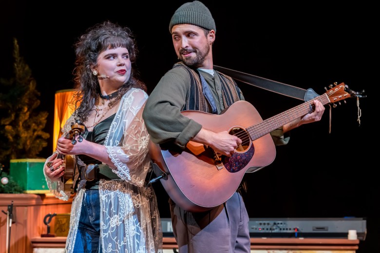 A close-up theatrical shot of two musicians. One person, wearing an earth-toned outfit, is singing and playing a large acoustic guitar, while the other person, in dark jeans and a sheer top, holds a small fiddle or violin and looks off to the side.