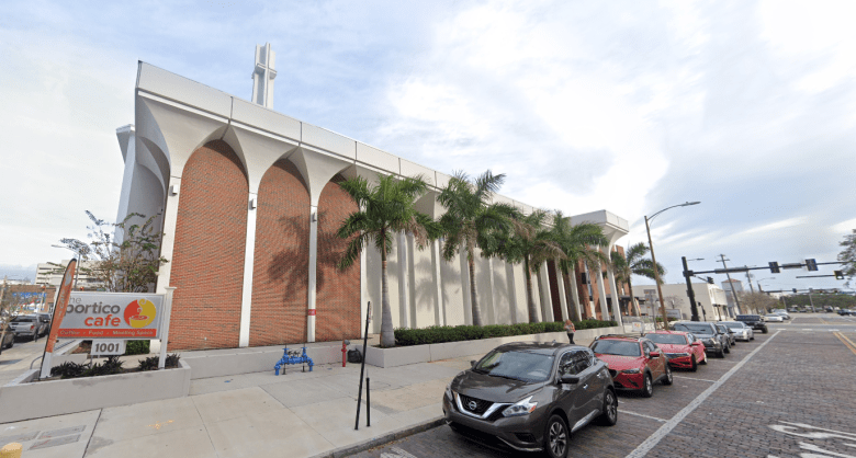 A wide-angle street-level view of a modern mid-century building with distinctive white concrete arches and red brick infill panels. A sign in the foreground identifies the location as "the portico cafe," and a row of palm trees is planted along the sidewalk.