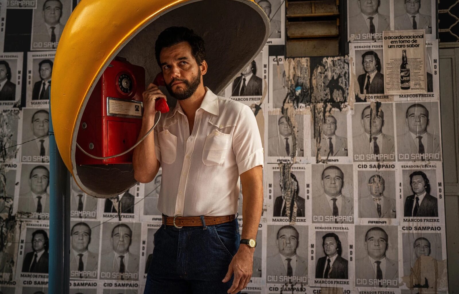 Photo of a man in a yellow, old-school telephone booth speaking on a red telephone. Behind him are black-and-white flyers with headshots plastered on a wall