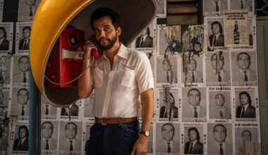 Photo of a man in a yellow, old-school telephone booth speaking on a red telephone. Behind him are black-and-white flyers with headshots plastered on a wall