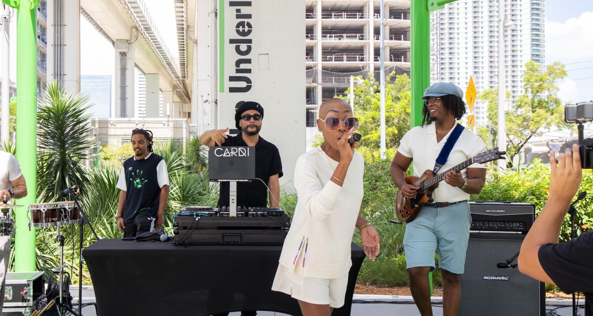 Photo of a bald woman on stage holding a microphone at an outdoors venue.