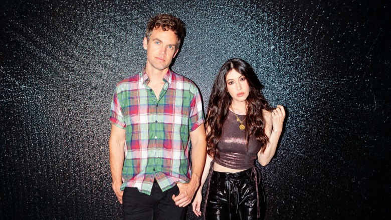: Two musicians posing for a photo. The subject on the left stands with their hand in their pocket, and the subject on the right is touching their long, dark, wavy hair, both looking directly at the camera with serious expressions.