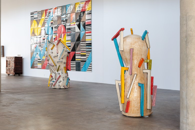 Two tall, abstract, beige sculptures with colorful geometric protrusions stand on a concrete floor in front of a large, colorful, tiled geometric wall artwork in a bright gallery space.