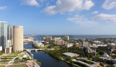 Aerial view of downtown Tampa showing the Hillsborough River, Riverwalk, high-rise office towers and neighborhoods stretching toward Tampa Bay under a bright blue sky.