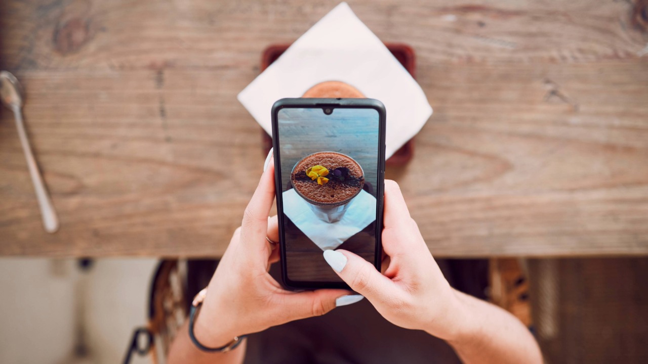 A person photographs a plated dessert with a smartphone at a café table, illustrating how food content is created for social media.