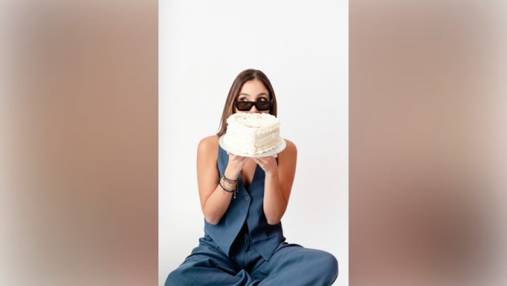 A woman holds a frosted cake in front of her face while wearing sunglasses, photographed against a white background.