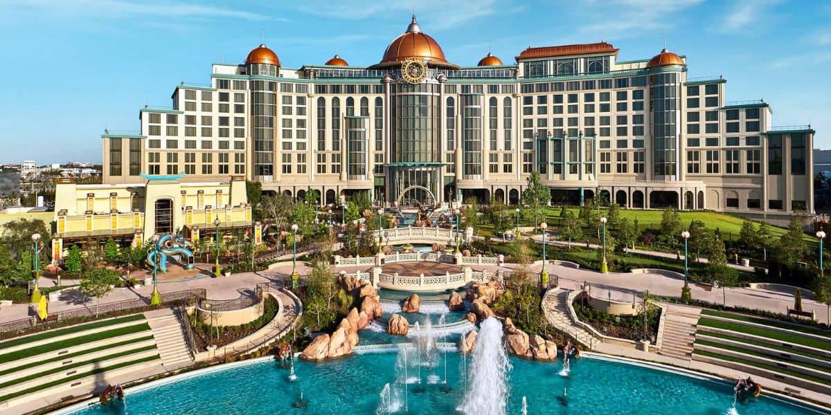 The grand, multi-story Grand Helios Hotel with ornate domes and a clock on top, surrounded by lush gardens, pathways, and a large fountain pool in the foreground under a clear blue sky. Located within Universal Orlando Resort's Epic Universe. Universal Epic Universe news coming out.